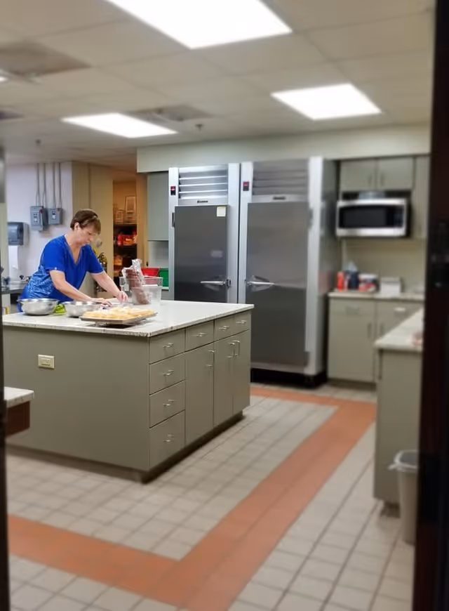 A woman in a blue shirt preparing food on a large kitchen island in a commercial kitchen. The kitchen has stainless steel refrigerators, a microwave, cabinets, and tiled flooring with a red border pattern.