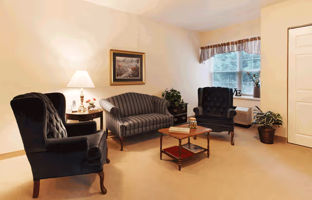 A cozy living room with two dark upholstered armchairs and a striped loveseat arranged around a wooden coffee table. A side table with a lamp and decorative items is next to one armchair. There is a framed painting on the wall, a window with a valance, and potted plants near the window and door.