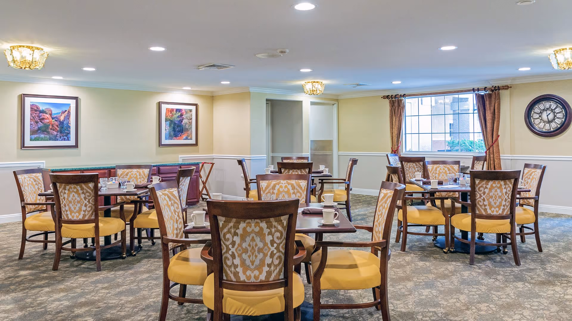 A dining room with multiple wooden tables and chairs with yellow cushions and patterned backs. Each table is set with white cups and saucers. The room has beige walls with white wainscoting, two framed landscape paintings, a large window with brown curtains, a wall clock, and ceiling lights.