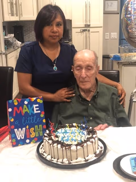 An elderly man sitting at a table with a birthday cake in front of him and a woman standing behind him with her hand on his shoulder in a kitchen setting. A colorful sign on the table reads 'MAKE a little WISH'.