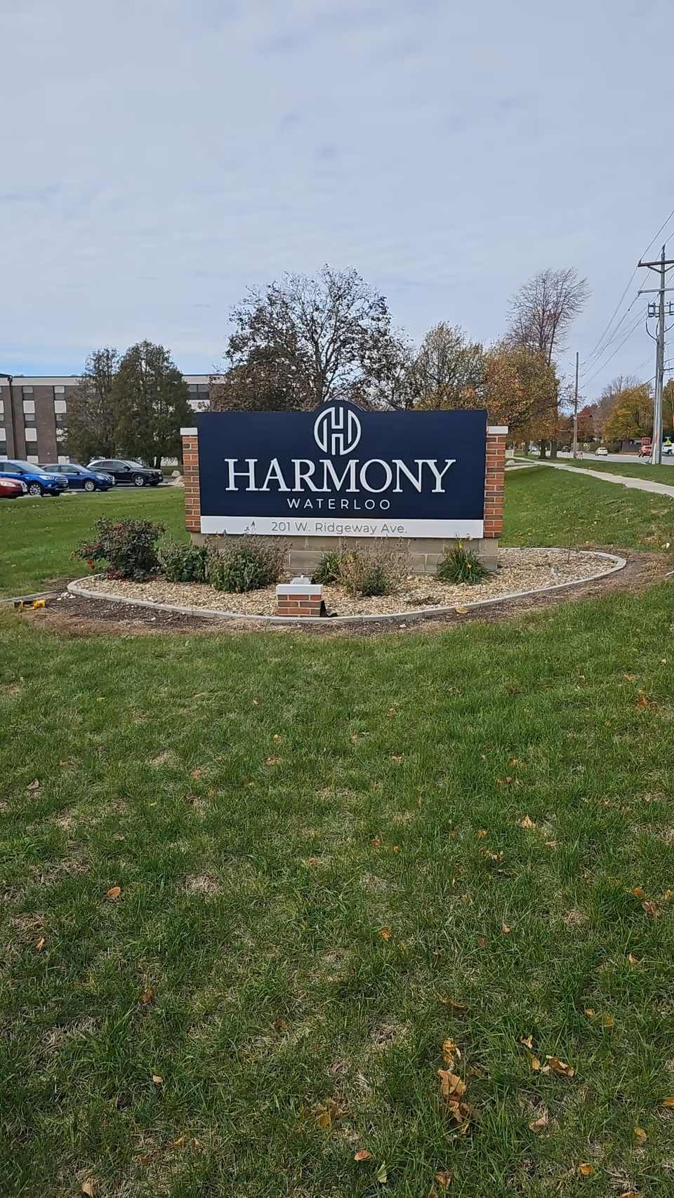 Freestanding sign reading "Harmony Waterloo" on a landscaped lawn outside the facility with a building and cars in the background.