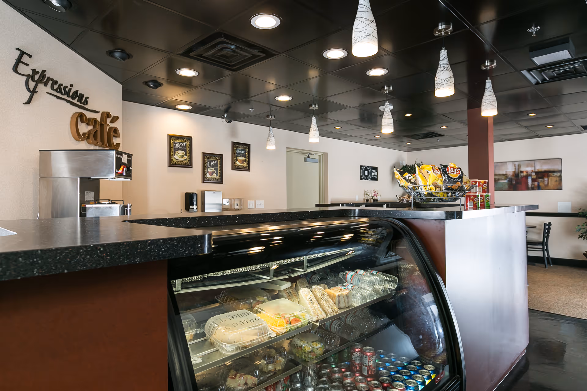 Interior café counter with a glass refrigerated display case stocked with drinks and sandwiches, a countertop holding chips and snacks, and an 'Expressions café' wall sign.