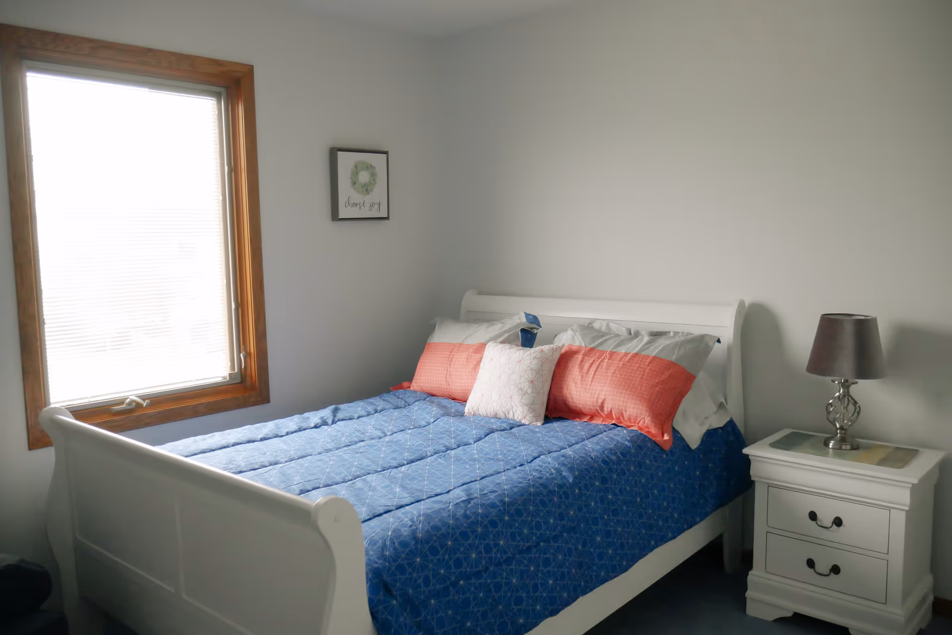 A bedroom with a white wooden bed frame, blue patterned bedspread, and several pillows in white and coral colors. There is a window with blinds on the left wall and a small framed picture hanging above the bed. A white nightstand with two drawers and a silver lamp with a dark lampshade is on the right side of the bed.