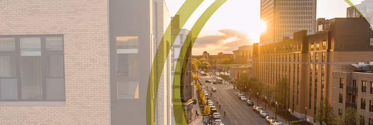 View down a sunlit urban street at sunset with brick buildings, parked cars, and a green translucent curved graphic overlay.