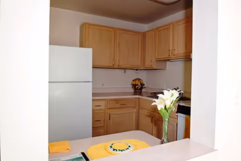 Small kitchen area with light wood cabinets, a white refrigerator, a stove, and a countertop. A vase with white flowers is placed on the counter, along with a yellow placemat and a folded yellow towel.