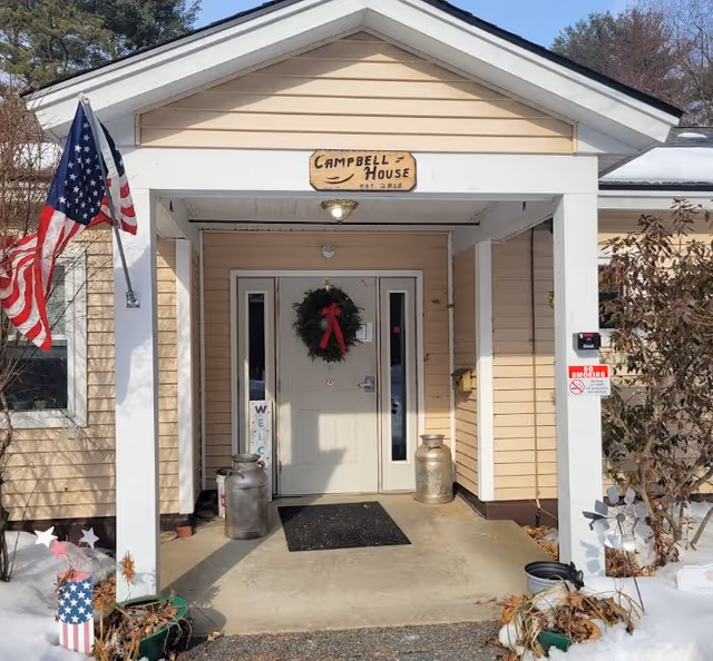 Front entrance of a beige house with white trim, decorated with a holiday wreath on the door and an American flag on the left side. There are two metal milk cans on either side of the door, a black doormat, and some potted plants with snow around the entrance. A wooden sign above the door reads 'Campbell House EST. 2016'.