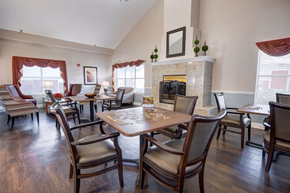 A bright and spacious common area with multiple tables and chairs arranged for social activities. One table in the foreground has a partially completed jigsaw puzzle. The room features a fireplace with decorative greenery on the mantel, large windows with red valances, and comfortable armchairs along the walls.