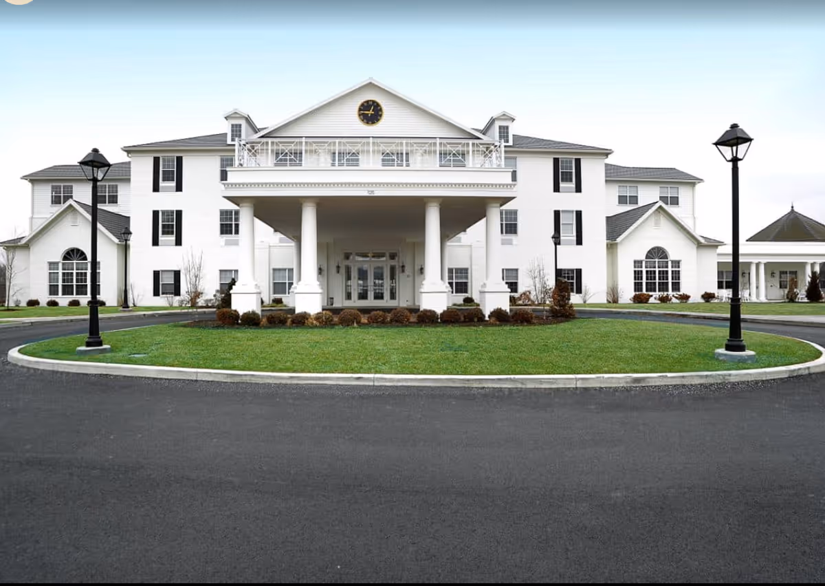 Front exterior view of a large white senior living facility building with a covered entrance supported by four columns, a clock on the top center of the building, black window shutters, and a circular driveway with green grass and two black lamp posts.