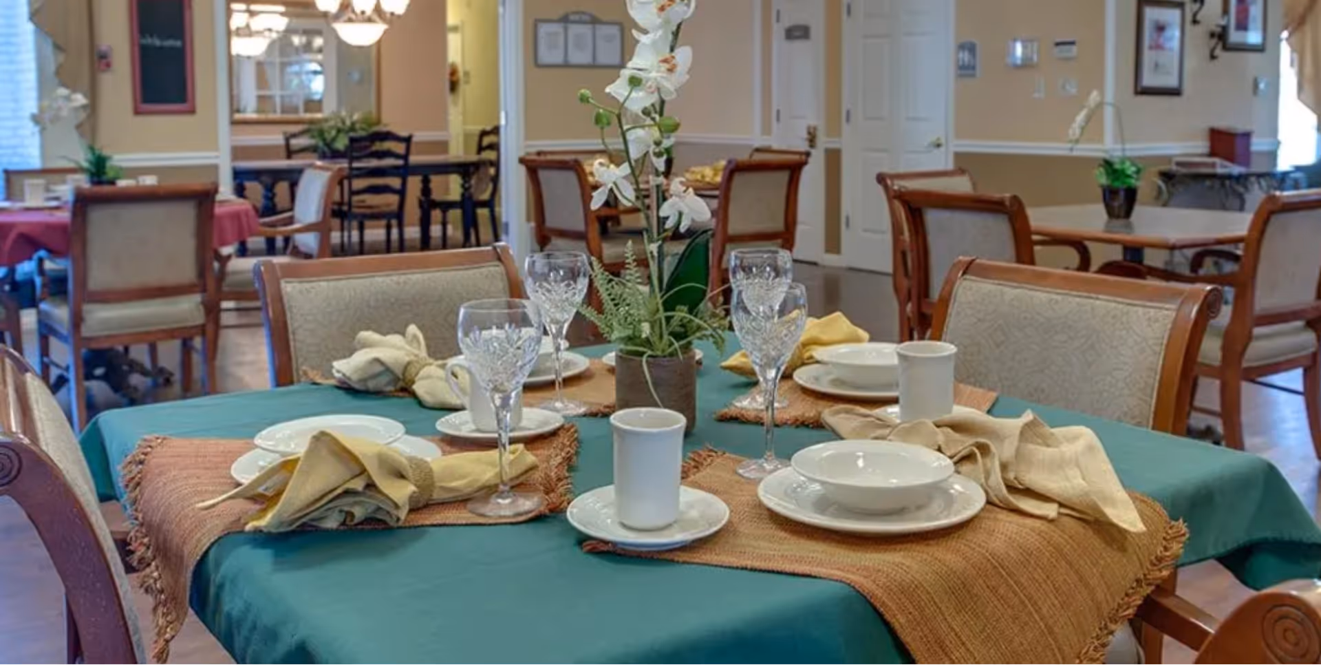 Dining room table set with plates, crystal glasses, cloth napkins and an orchid centerpiece in a senior living facility dining area.