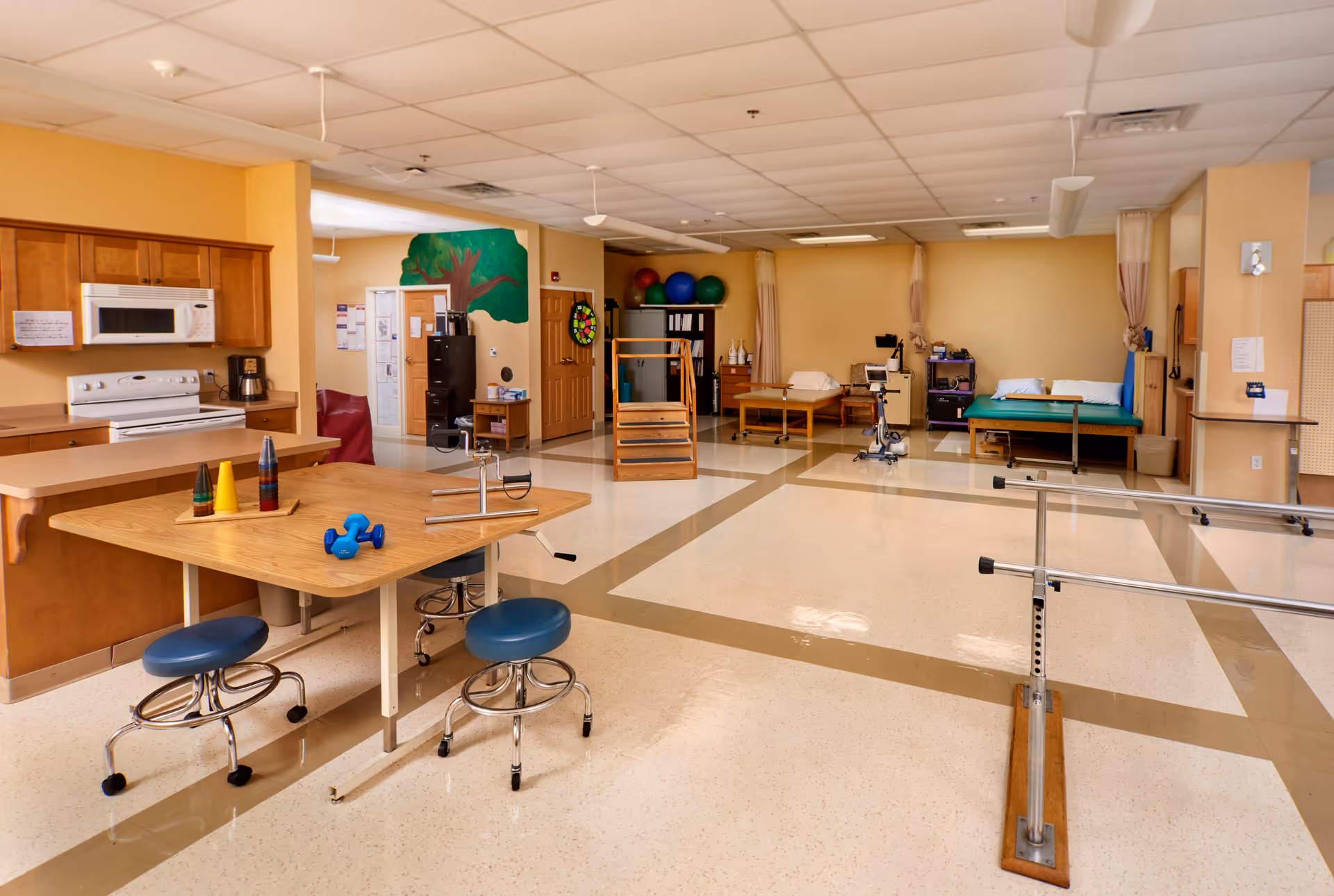 A spacious rehabilitation or physical therapy room with exercise equipment including parallel bars, therapy tables, exercise balls, and small weights. There is a kitchen area with a microwave, stove, and coffee maker on the left side. The room has beige walls and a tiled floor with a grid pattern.