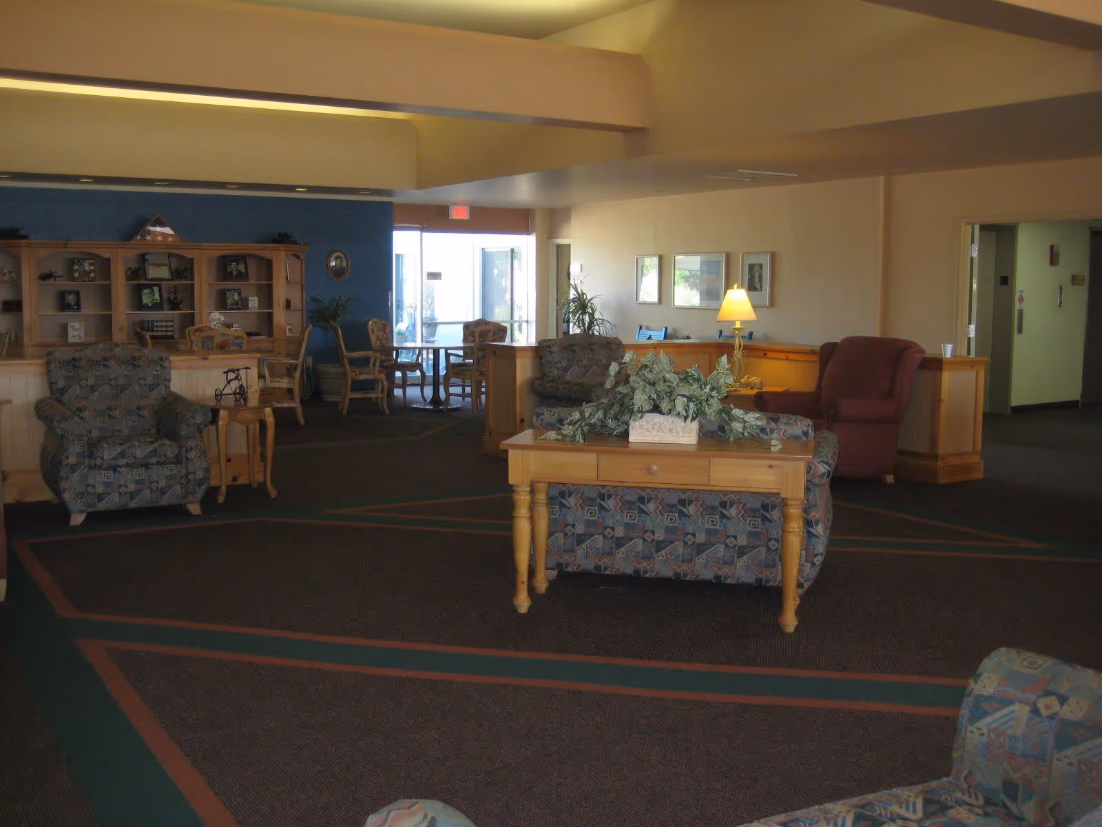 A spacious senior living facility common area with patterned armchairs, a wooden table with a plant centerpiece, a lamp, and a bookshelf with framed photos and decorations. There is a seating area with chairs and tables near a glass door entrance.