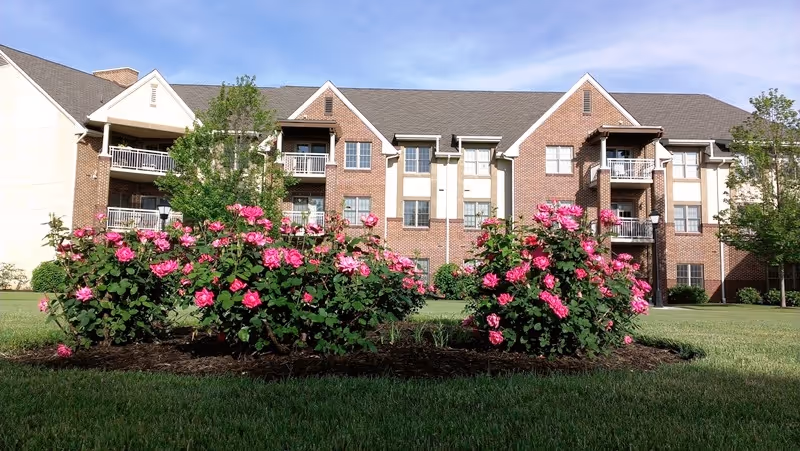A three-story brick residential building with balconies, surrounded by a well-maintained lawn and vibrant pink rose bushes in the foreground under a clear blue sky.
