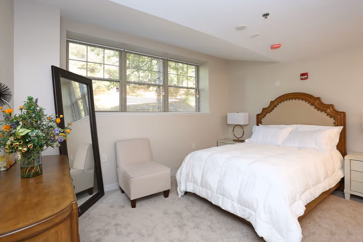 Neutral-toned bedroom with a white-made bed and wooden headboard, window, armchair, leaning full-length mirror, and a dresser with flowers.