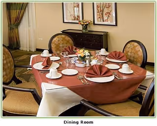 A dining room table set for six with a red tablecloth and white undercloth, each place setting includes a white plate, cup, saucer, silverware, and a folded red napkin. The table has a centerpiece with flowers and a bowl of snacks. Four upholstered chairs surround the table, and there is a dark wooden sideboard with a flower vase against the wall. Two framed floral artworks hang above the sideboard.