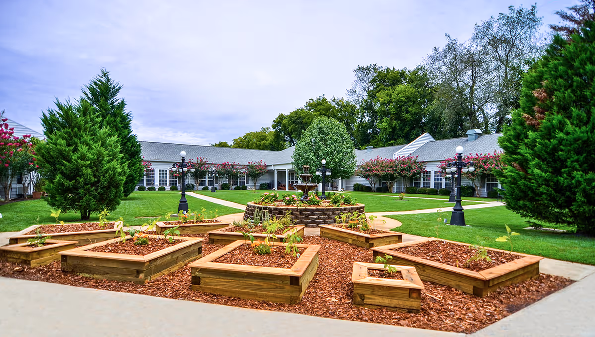 Outdoor garden area at The Neighborhood at Sheffield featuring multiple raised wooden garden beds with small plants, a circular brick planter with a fountain in the center, green grass, trees, and a white building with many windows in the background under a cloudy sky.