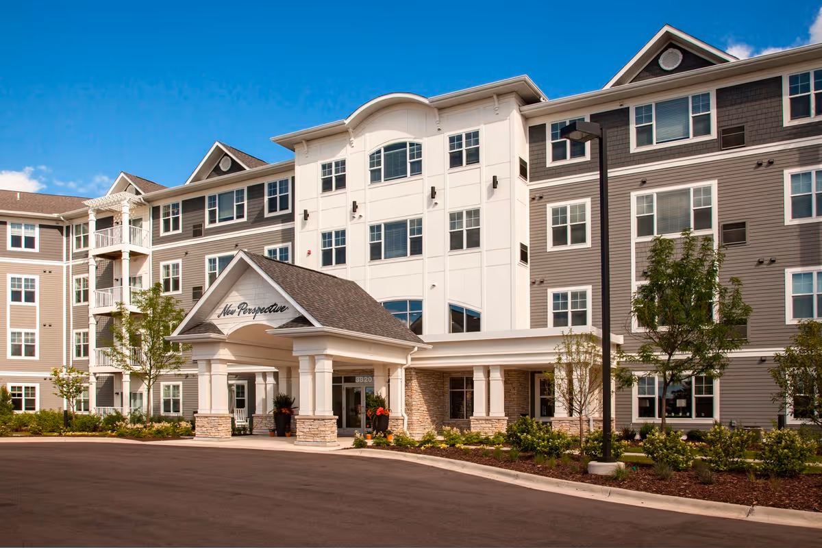Exterior view of a multi-story senior living facility named New Perspective Senior Living | White Bear Lake, featuring a covered entrance with columns, landscaped greenery, and a clear blue sky.
