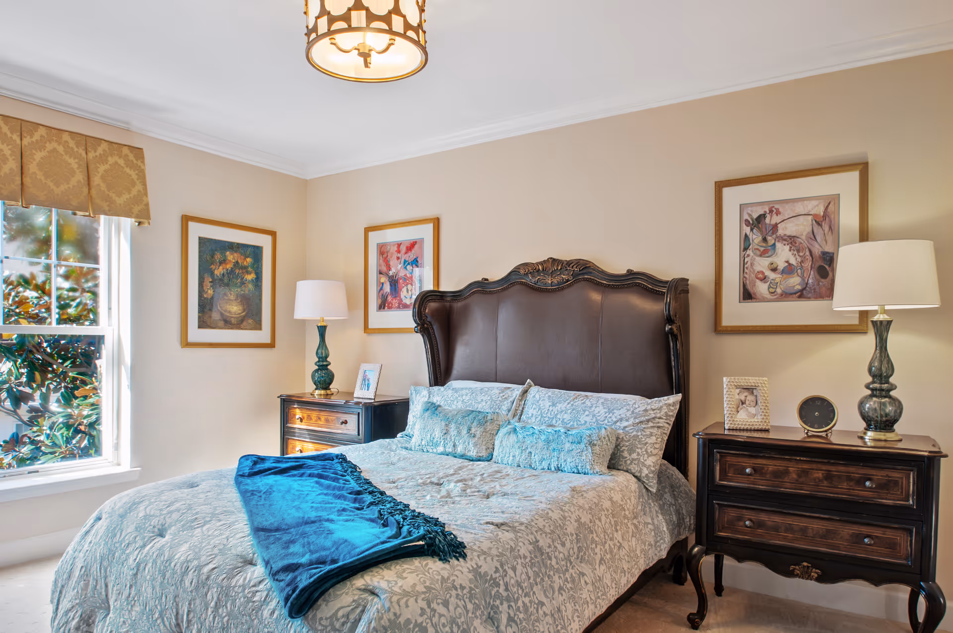 A well-decorated bedroom with a large dark wooden headboard bed covered in light blue patterned bedding and a dark blue throw blanket. There are two matching dark wooden nightstands on either side of the bed, each with a lamp and decorative items. The walls are beige with three framed paintings hanging above the bed. A window with a patterned valance lets in natural light from the left side.