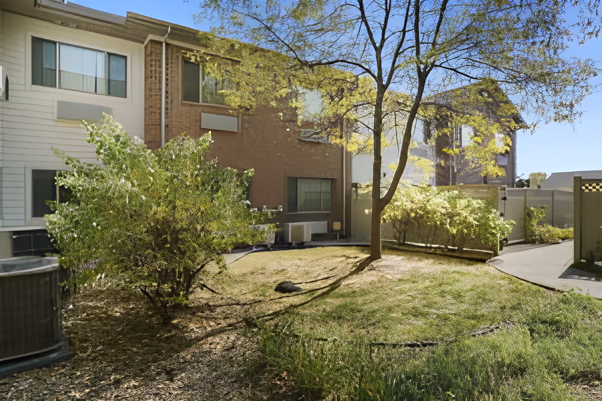 Outdoor view of a senior living facility courtyard with green grass, bushes, and trees. The building exterior is visible with windows and a pathway leading through the area under a clear blue sky.