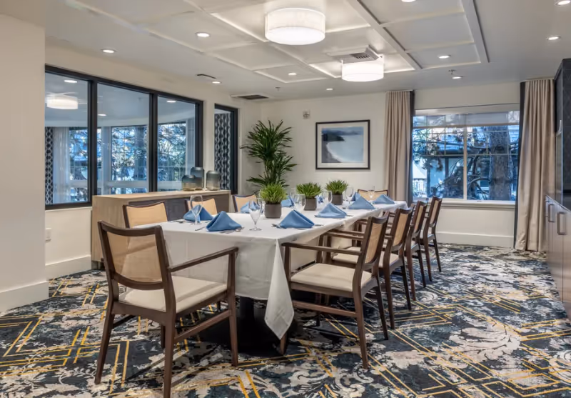 A well-lit dining room with a long rectangular table covered with a white tablecloth, set with blue folded napkins, glassware, and small potted plants as centerpieces. The room features patterned carpet, large windows with beige curtains, modern ceiling lights, and wooden chairs with cushioned seats around the table.
