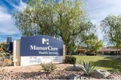Entrance sign for ManorCare Health Services amid landscaped grounds with trees and the facility building visible in the background.