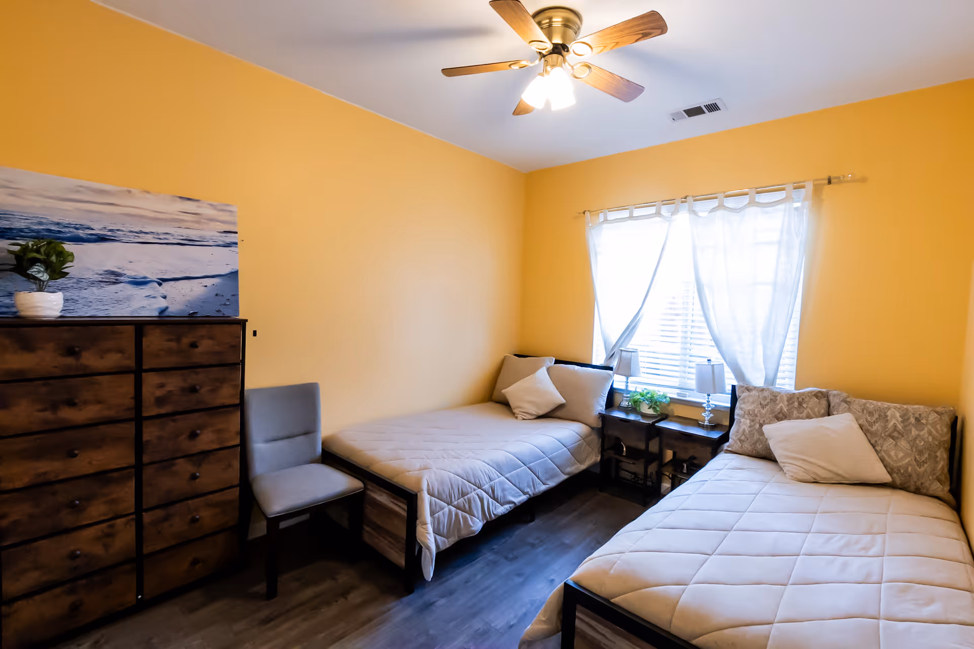 A bedroom with two single beds, each with pillows and beige bedding. Between the beds is a small nightstand with two lamps and a small plant. A window with sheer white curtains allows natural light into the room. On the left side, there is a wooden dresser with a potted plant and a landscape picture on top. A gray chair is placed next to the dresser. The walls are painted yellow, and a ceiling fan with lights is mounted on the ceiling.