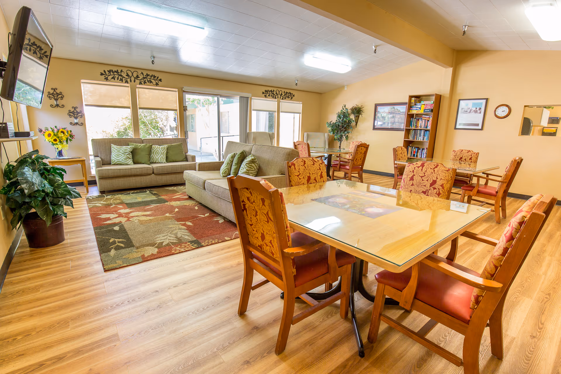 A bright and spacious common area with wooden flooring, featuring a glass-top table surrounded by six upholstered chairs with floral patterns. In the background, there are two beige sofas with green cushions placed on a patterned area rug. Large windows with beige blinds allow natural light to fill the room. The walls are decorated with framed pictures, a clock, and a bookshelf filled with books and board games. Several potted plants add a touch of greenery to the space.