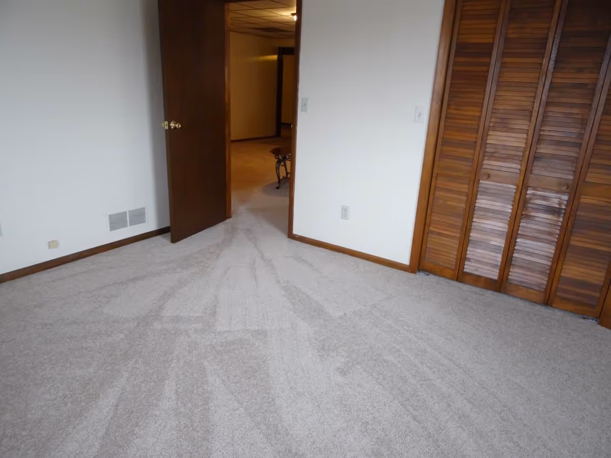 Empty carpeted room with wooden louvered closet doors and an open doorway to a hallway.