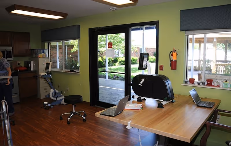 Bright multipurpose activity room with a large wooden table, exercise equipment, laptops, and glass doors opening to an outdoor courtyard.