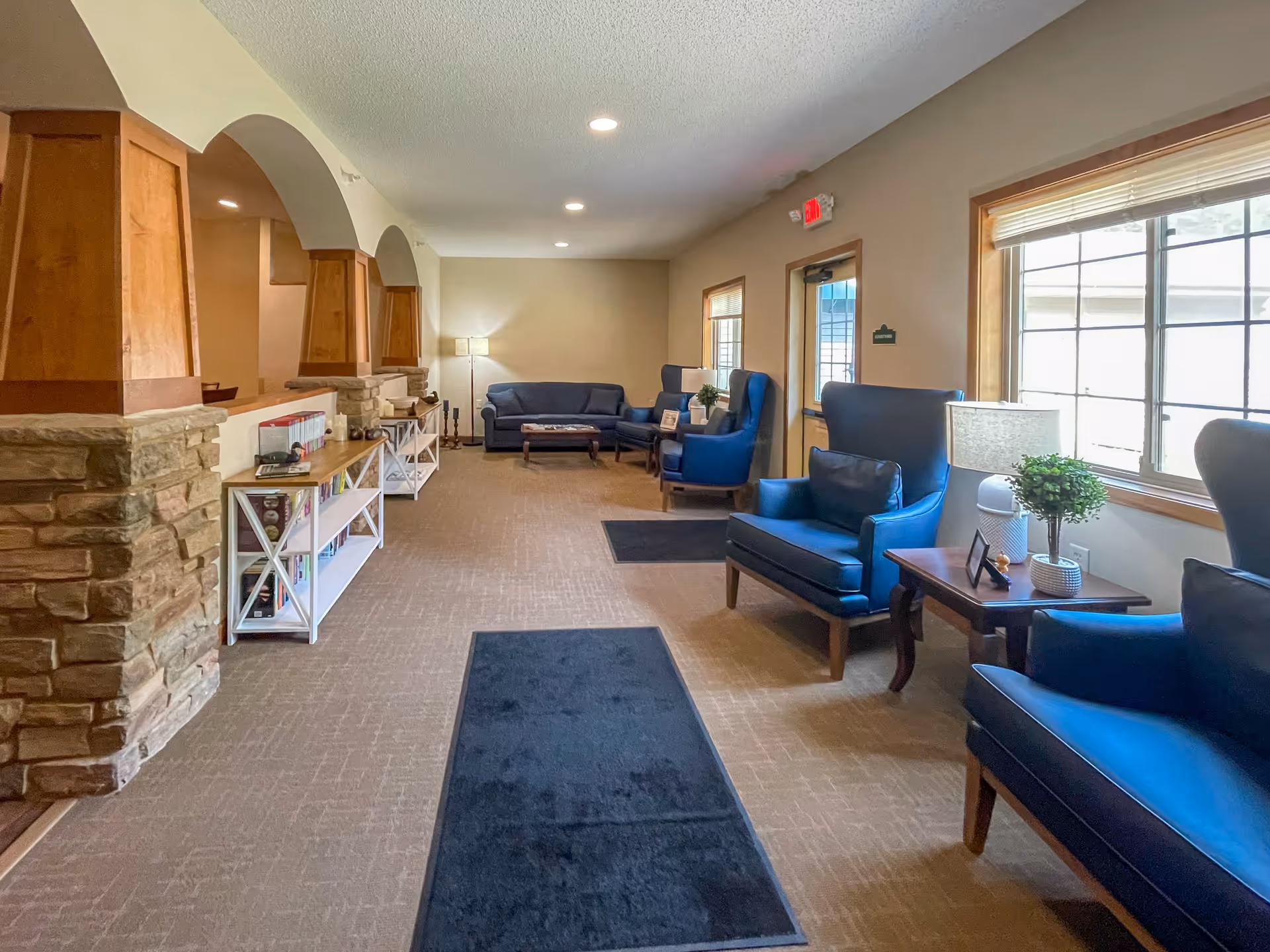 A well-lit lounge area with blue armchairs and sofas arranged along the walls. There are side tables with lamps and small plants, large windows letting in natural light, and a carpeted floor with black rugs. The space features stone and wood architectural details with arches and shelves holding books and decorative items.