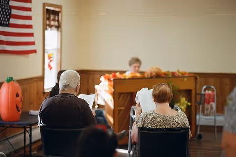 Two elderly individuals sitting in chairs facing a woman playing a piano in a room decorated with autumn and Halloween-themed decorations, including a large inflatable pumpkin and an American flag on the wall.