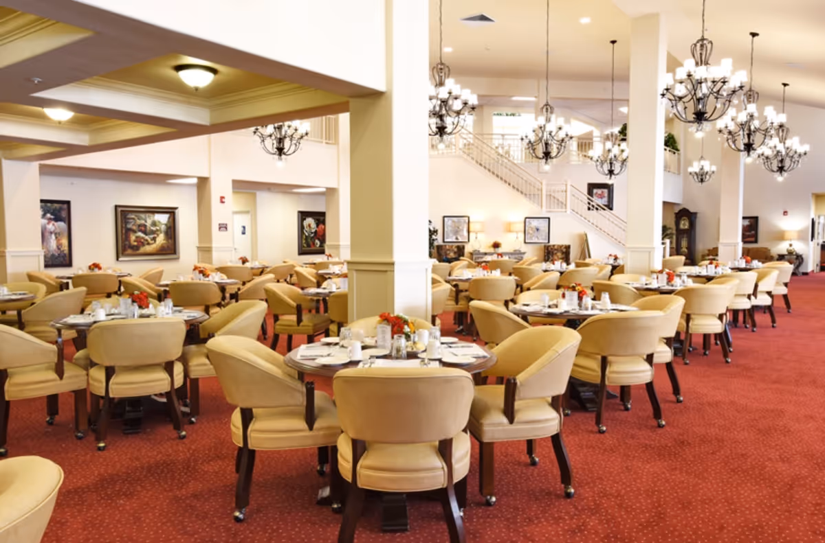 Spacious dining room with multiple round tables set with white plates, cups, and silverware. Beige cushioned chairs surround each table. The room features red carpet flooring, cream-colored walls, several chandeliers hanging from the ceiling, and framed artwork on the walls. A staircase and additional seating area are visible in the background.