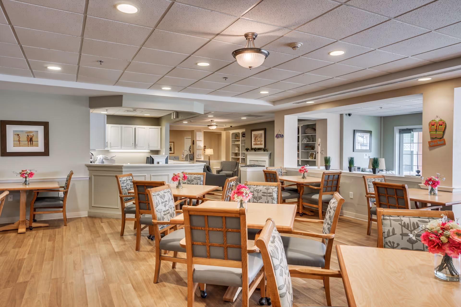 A bright and spacious dining area with several wooden tables and cushioned chairs arranged neatly. Each table has a small vase with pink flowers. The room features wood flooring, a white ceiling with recessed lighting, and light-colored walls. In the background, there is a kitchen area with white cabinets and a counter, and a cozy seating area with armchairs and a fireplace.