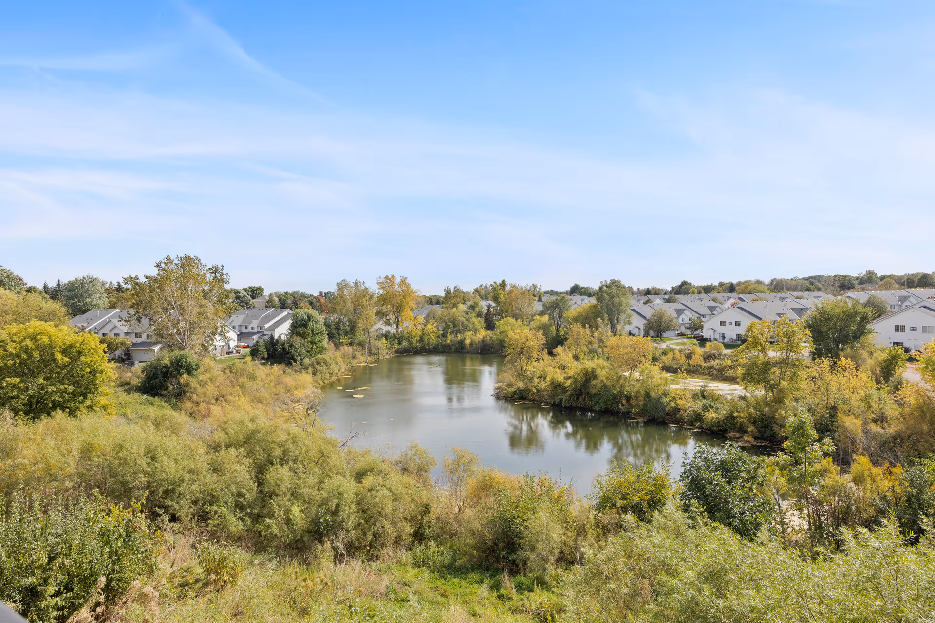 A scenic view of a small lake surrounded by dense green and yellow foliage with residential buildings in the background under a clear blue sky.