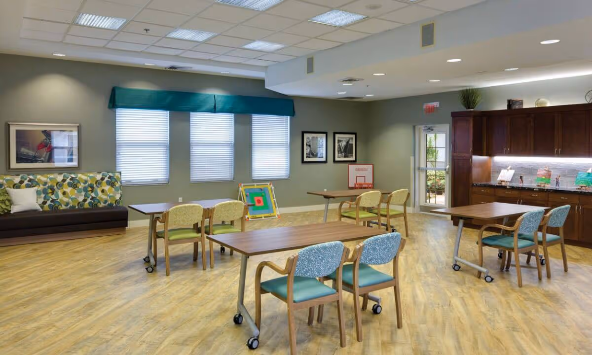 A spacious, well-lit common room with wooden tables and chairs on wheels, a cushioned bench with a colorful patterned backrest, and large windows with blinds and a teal valance. The room has light wood flooring, green walls, framed pictures, and a wooden cabinet with countertop and decorations. A door leads outside to a garden area.