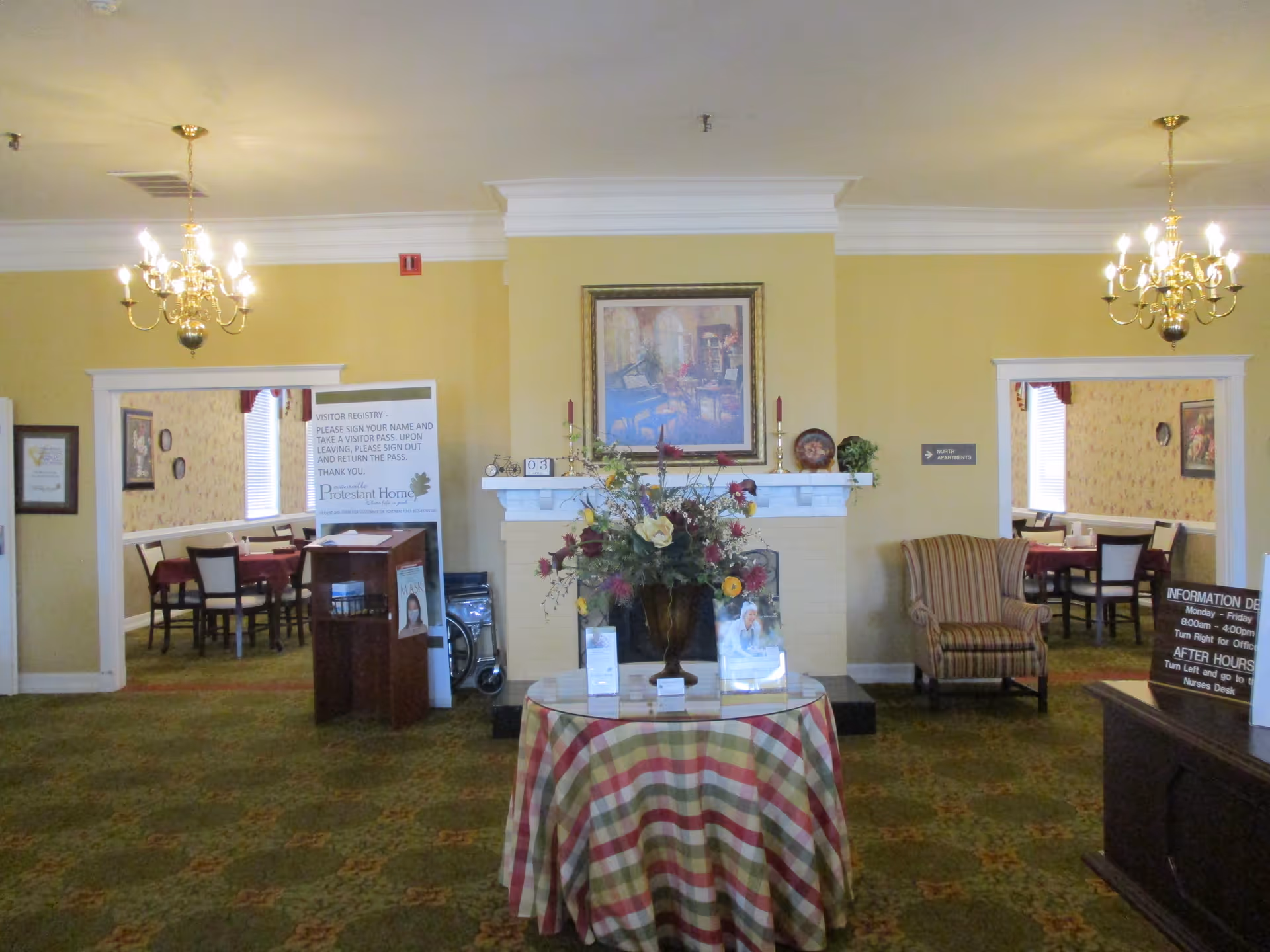 Interior view of a senior living facility lobby area with a round table covered with a checkered tablecloth and a flower arrangement on top. There is a fireplace with a framed painting above it in the center of the back wall. Two chandeliers hang from the ceiling. Two doorways on either side lead to dining areas with tables and chairs. A striped armchair is placed near the right doorway. Various informational signs and a visitor registry stand are visible.