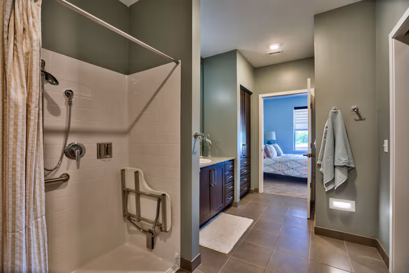 A spacious accessible bathroom showing a tiled walk-in shower with a fold-down seat, a vanity, and an open doorway to a bedroom.