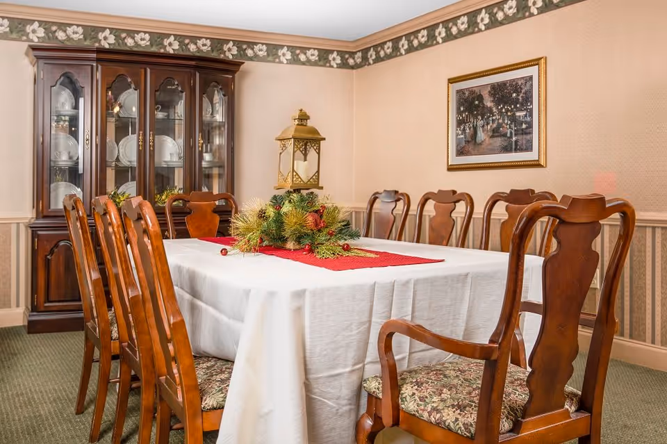 A formal dining room with a long rectangular table covered with a white tablecloth and a red runner. The table is surrounded by wooden chairs with floral cushioned seats. A decorative centerpiece with greenery and a large lantern is on the table. In the background, there is a wooden china cabinet displaying plates and glassware. The walls have a floral wallpaper border near the ceiling and a framed painting hangs on one wall.
