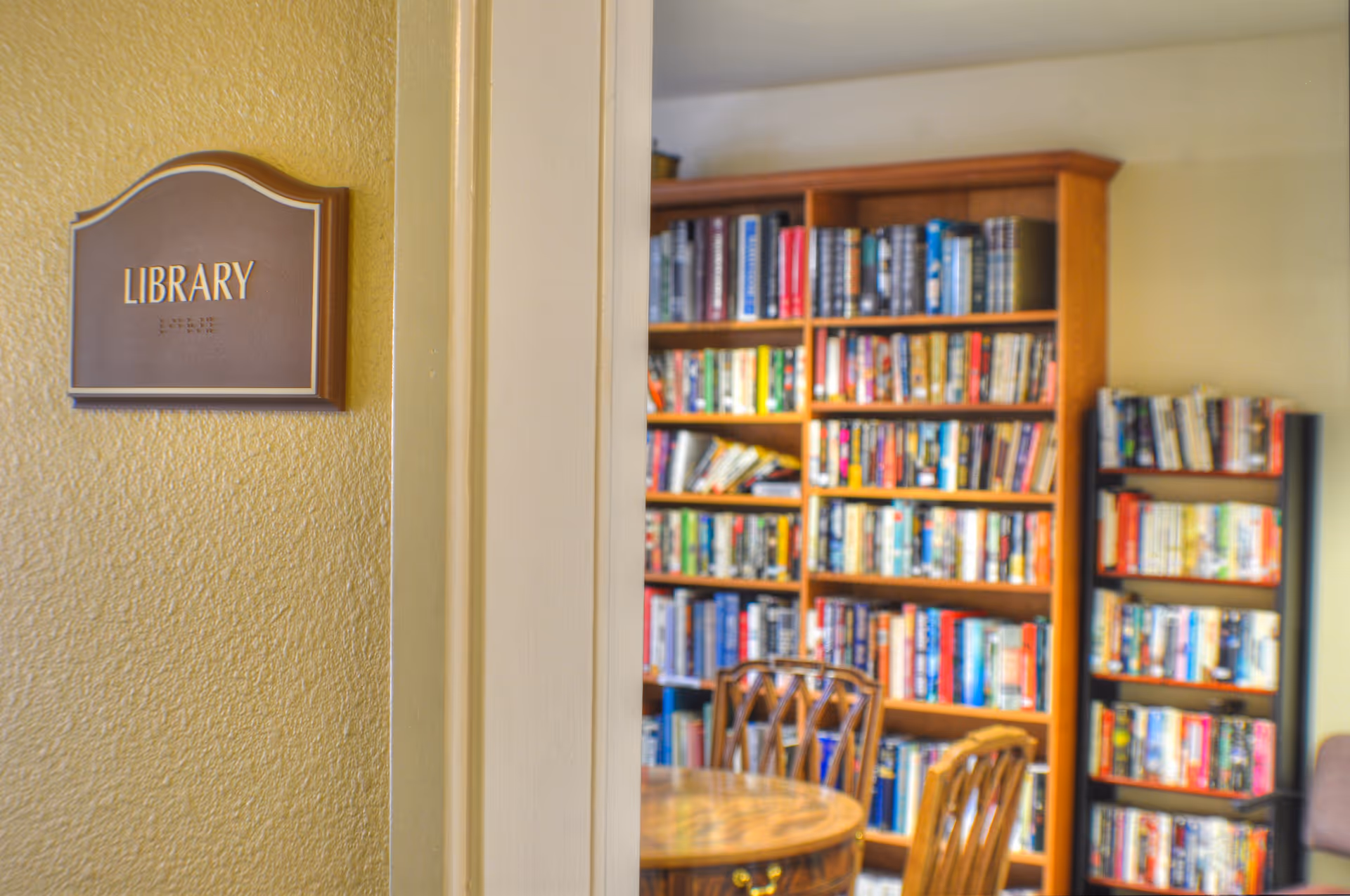 View of a library room in a senior living facility with a wooden sign labeled 'LIBRARY' on a textured yellow wall. Inside the room, there are wooden bookshelves filled with books and a round wooden table with chairs.