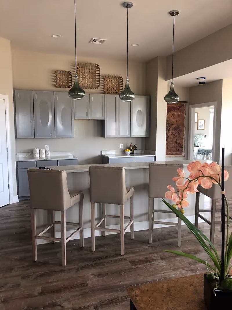 Open kitchen with a center island, three beige bar stools, hanging pendant lights and gray cabinets.