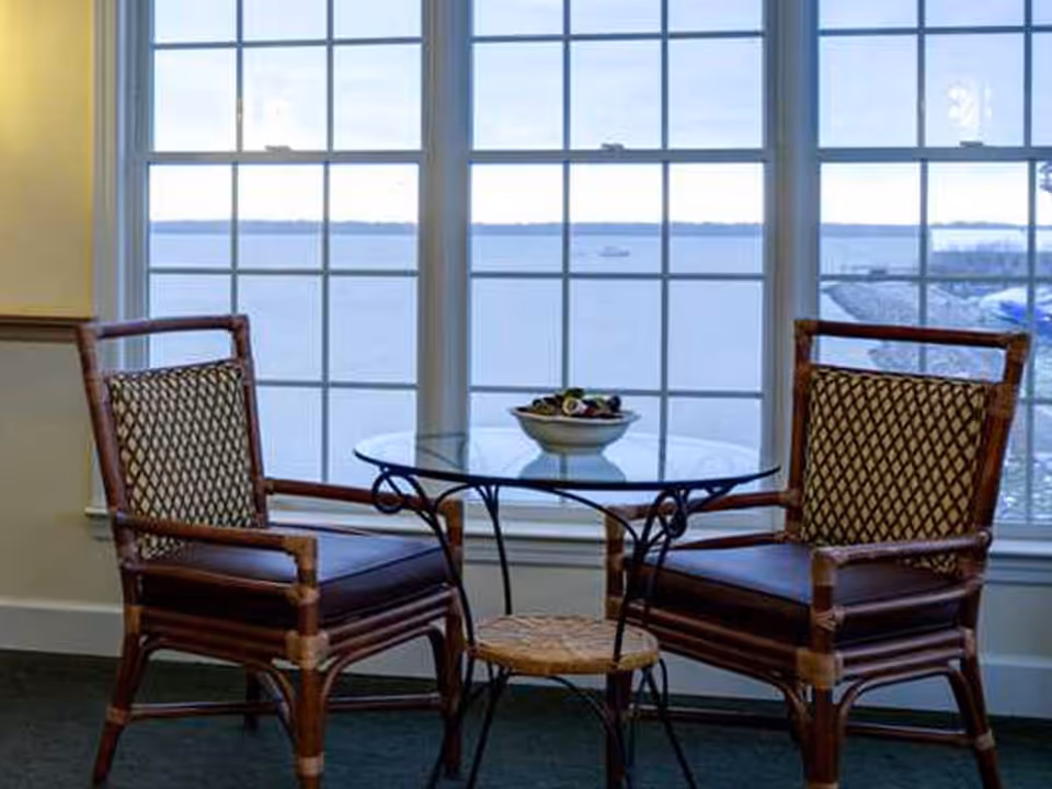 Two wooden chairs with patterned cushions positioned around a small round glass table with a decorative bowl on it, set in front of large windows overlooking a body of water and distant shoreline.