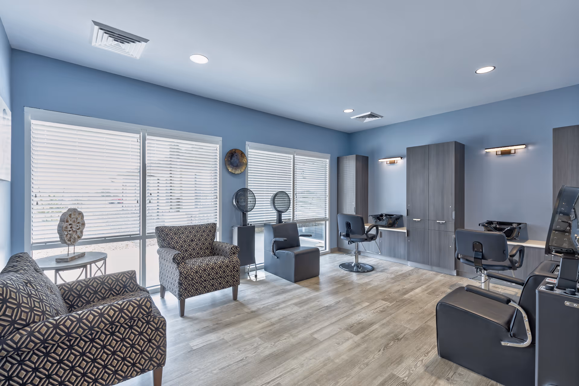 Bright salon-style room with styling chairs, hair-wash stations, patterned armchairs and large windows.