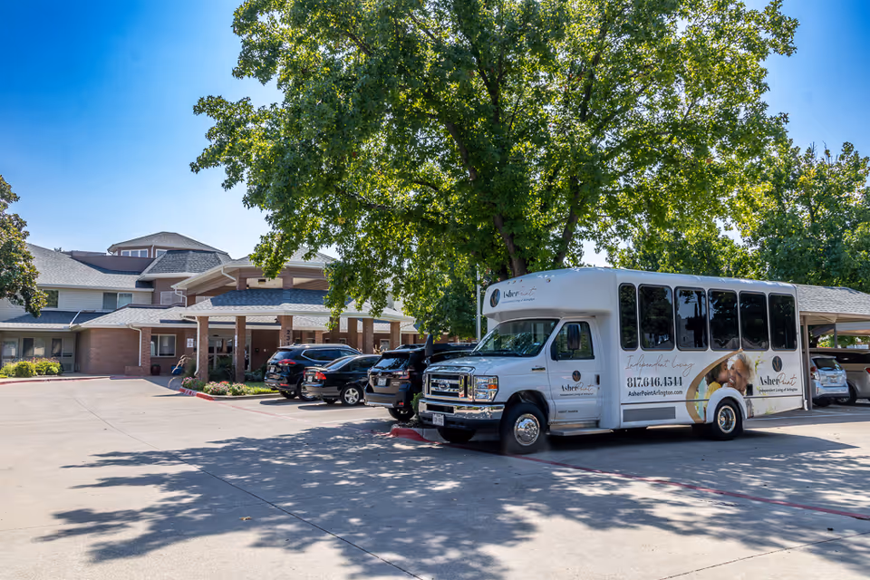 Front exterior of a senior living facility with a white shuttle bus parked under a large tree near the entrance.