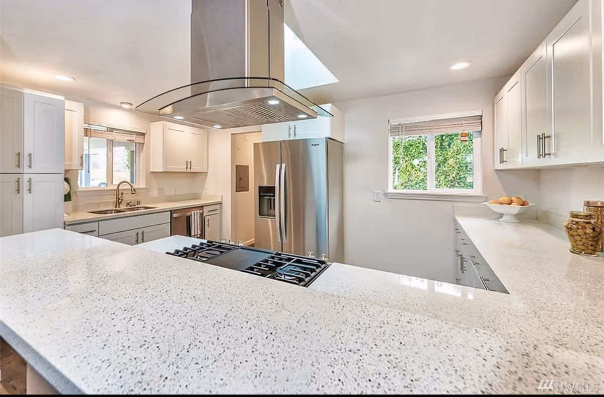 Modern kitchen with white cabinets, a large speckled white countertop, a stainless steel refrigerator, a gas stove with a stainless steel range hood, and two windows letting in natural light.