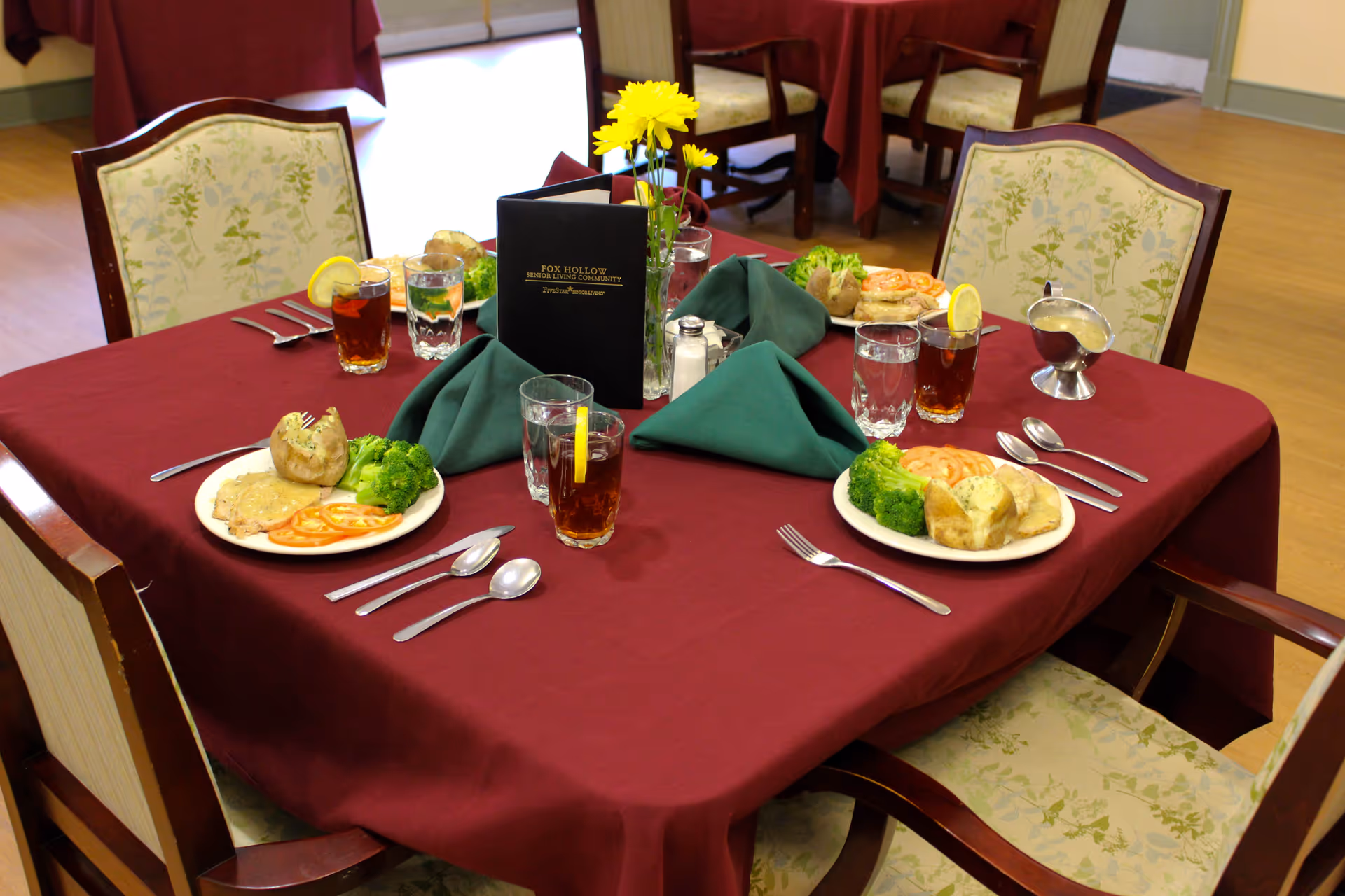 A dining table set for three with plates of food including baked potatoes, broccoli, and sliced tomatoes. Each place setting has a glass of iced tea with a lemon slice and a glass of water. The table is covered with a burgundy tablecloth and has green cloth napkins folded in the center along with a small vase of yellow flowers and a menu from Fox Hollow Senior Living Community.