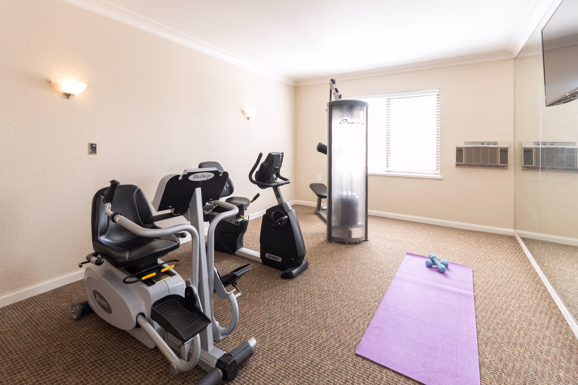 A small fitness room with exercise equipment including a recumbent bike, a stationary bike, and a multi-function weight machine. There is a purple yoga mat with two small dumbbells on the carpeted floor. The room has beige walls, a window with blinds, two wall-mounted air conditioning units, and a large mirror on one wall.