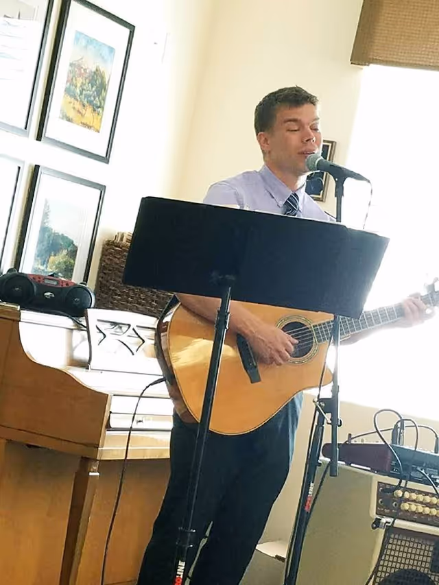 A man wearing a light purple shirt and tie is playing an acoustic guitar and singing into a microphone in a room with framed pictures on the wall and a piano in the background.