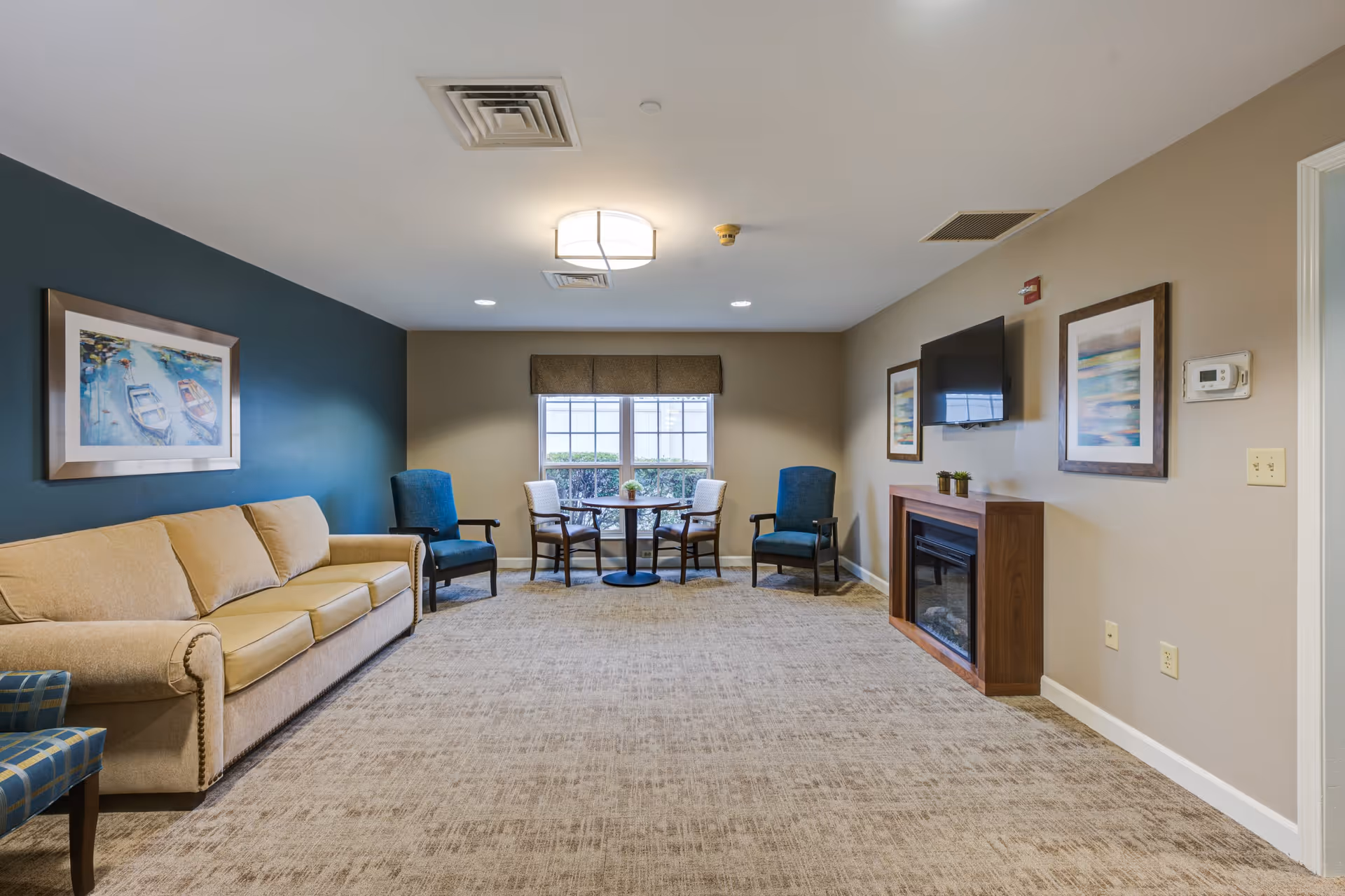 A cozy living room area in a senior living facility with a beige couch, two blue armchairs, a small round table with four chairs near a window, a wall-mounted TV above a wooden electric fireplace, and framed artwork on the walls.