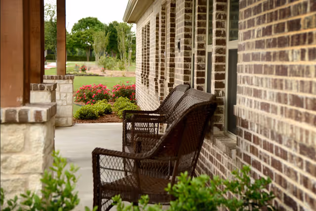 A cozy porch area featuring brown wicker chairs beside a brick wall, with colorful flowers and greenery in the background.