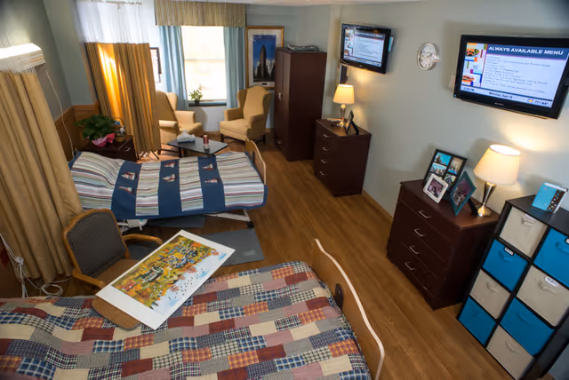 A well-lit senior living facility room with two beds covered in patterned quilts. One bed has a colorful puzzle on it. The room features wooden flooring, two armchairs near a window with curtains, a wooden dresser with framed photos and a lamp, a cabinet, and two wall-mounted televisions displaying menus and information. The walls are light-colored, and there is a storage unit with blue and beige fabric bins.