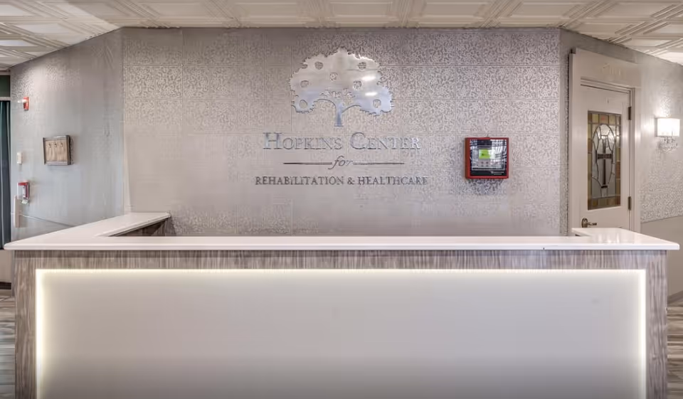 Reception desk area inside Hopkins Center for Rehabilitation and Healthcare with a decorative wall featuring the facility's name and logo, a fire alarm box, and a door with stained glass on the right.
